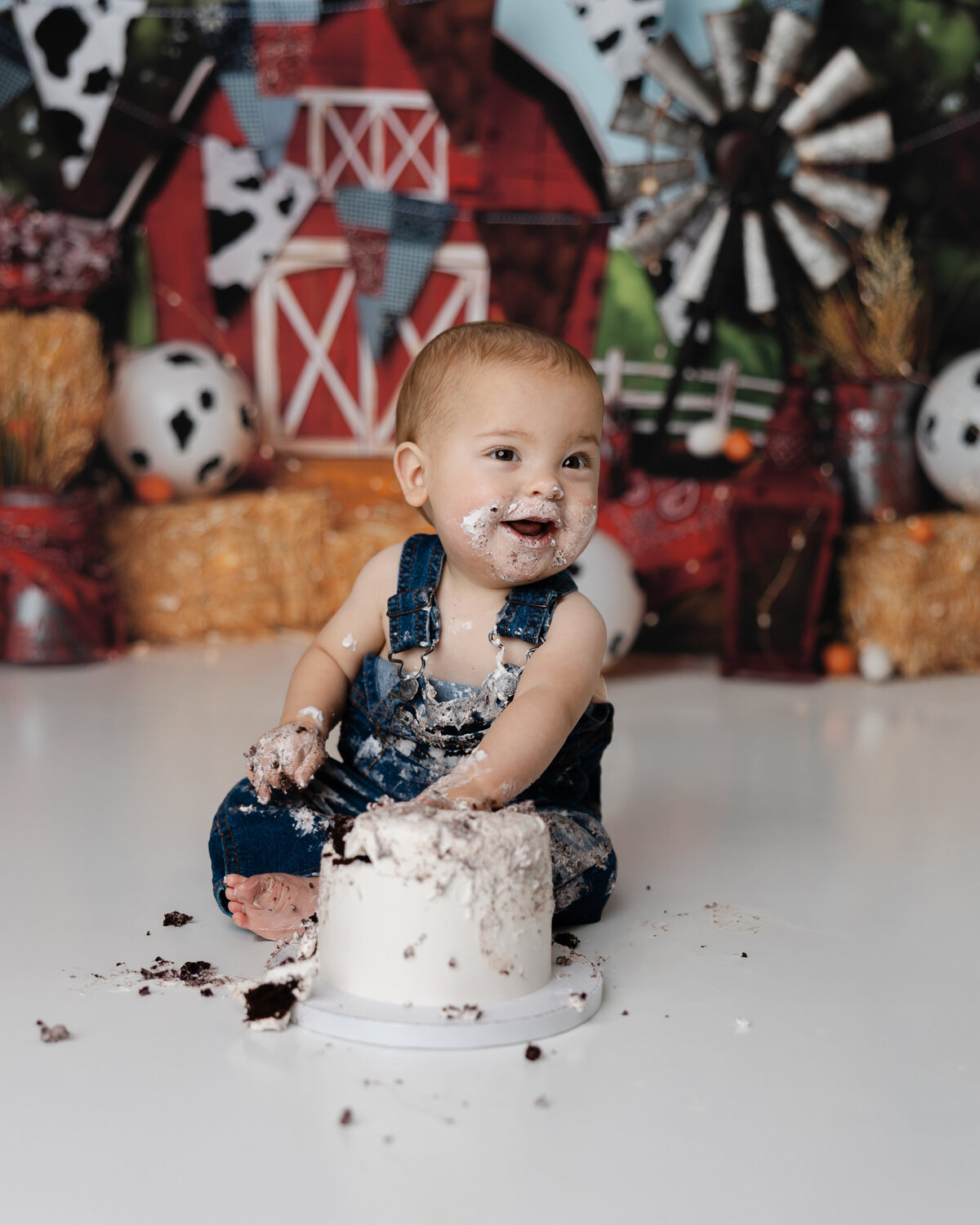 Laughing baby in overalls enjoying his cake smash in front of a red barn backdrop decorated with cow balloons and rustic props. Photograph by Yucaipa cake smash photographer Kaitlyn Dawn Photography.