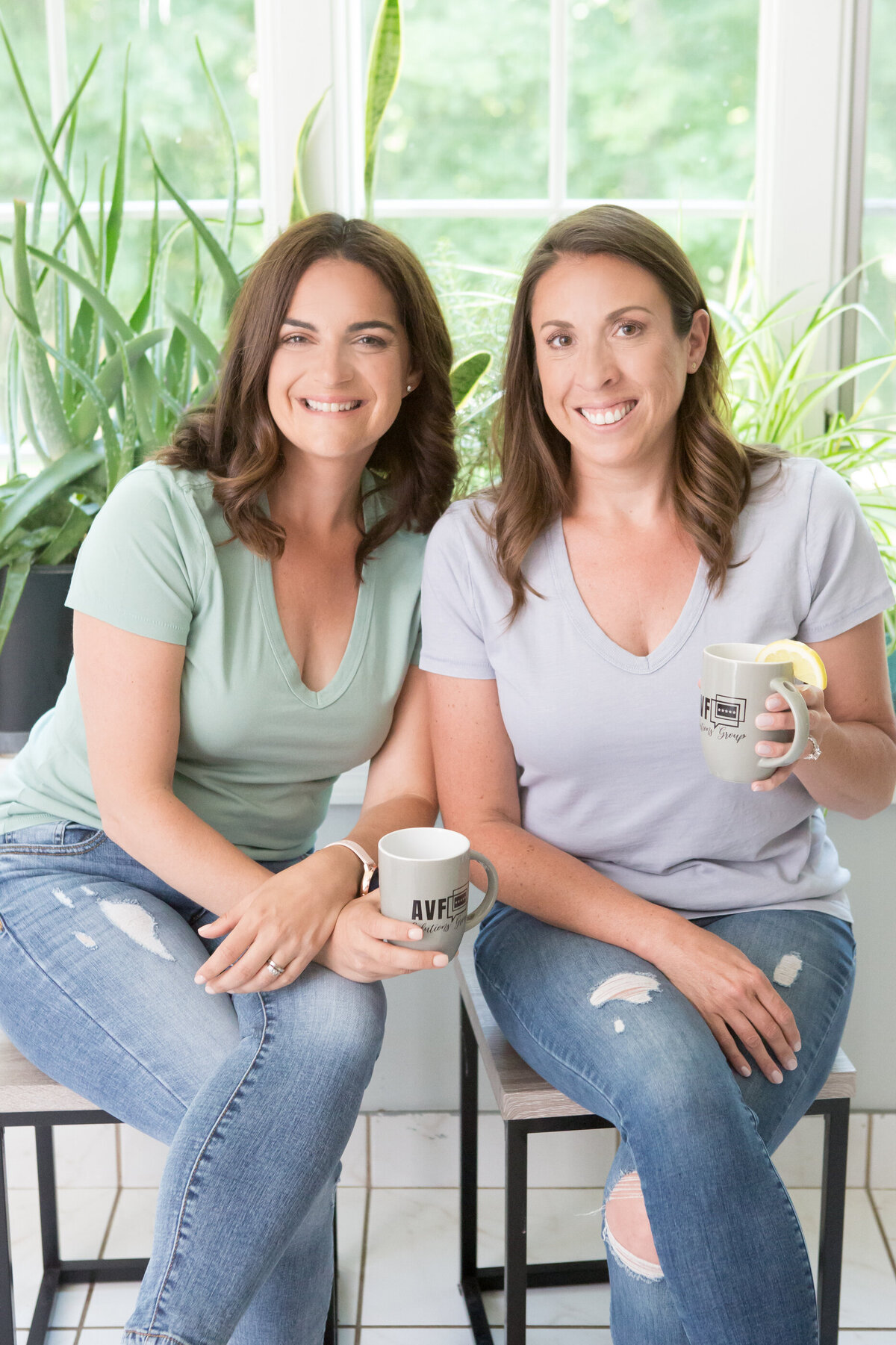 Two white women in a t-shirts and blue jeans hold mugs and smile in front of a bright window with house plants New York branding photographer Milestone Images