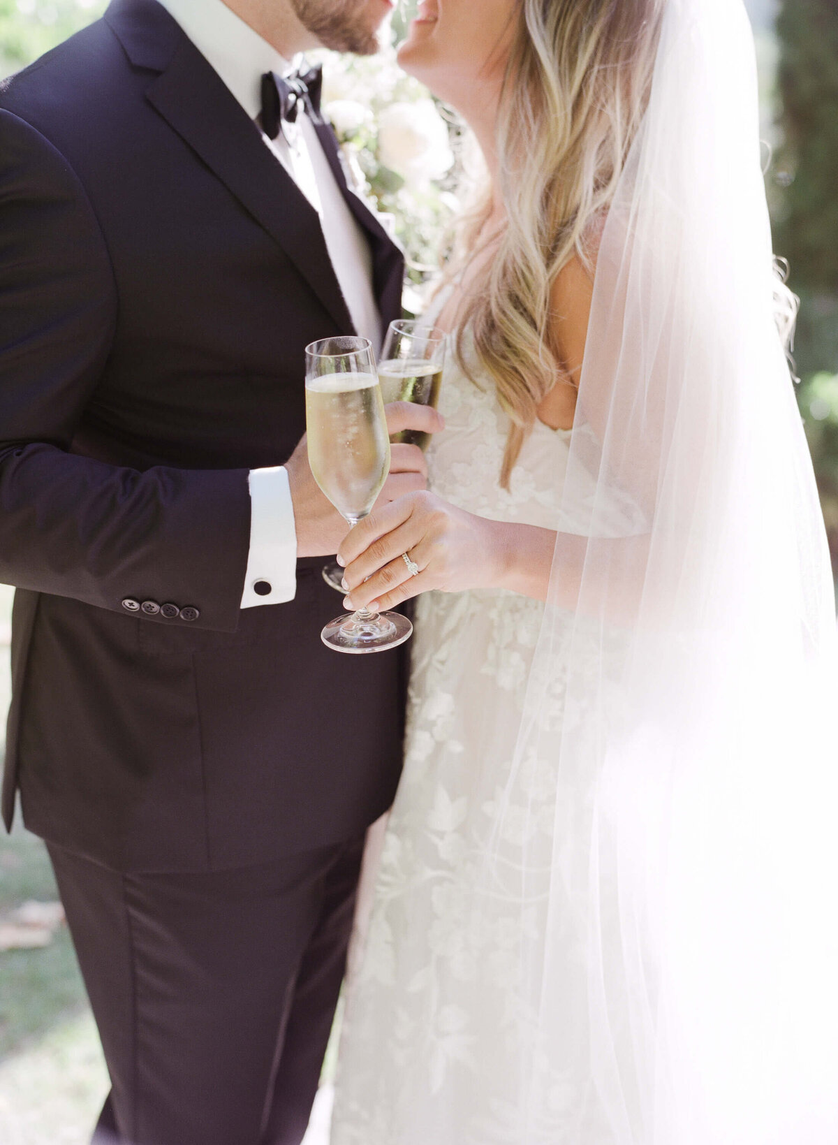 Happily married bride and groom lean into each other for a kiss while holding fizzy champagne glasses.