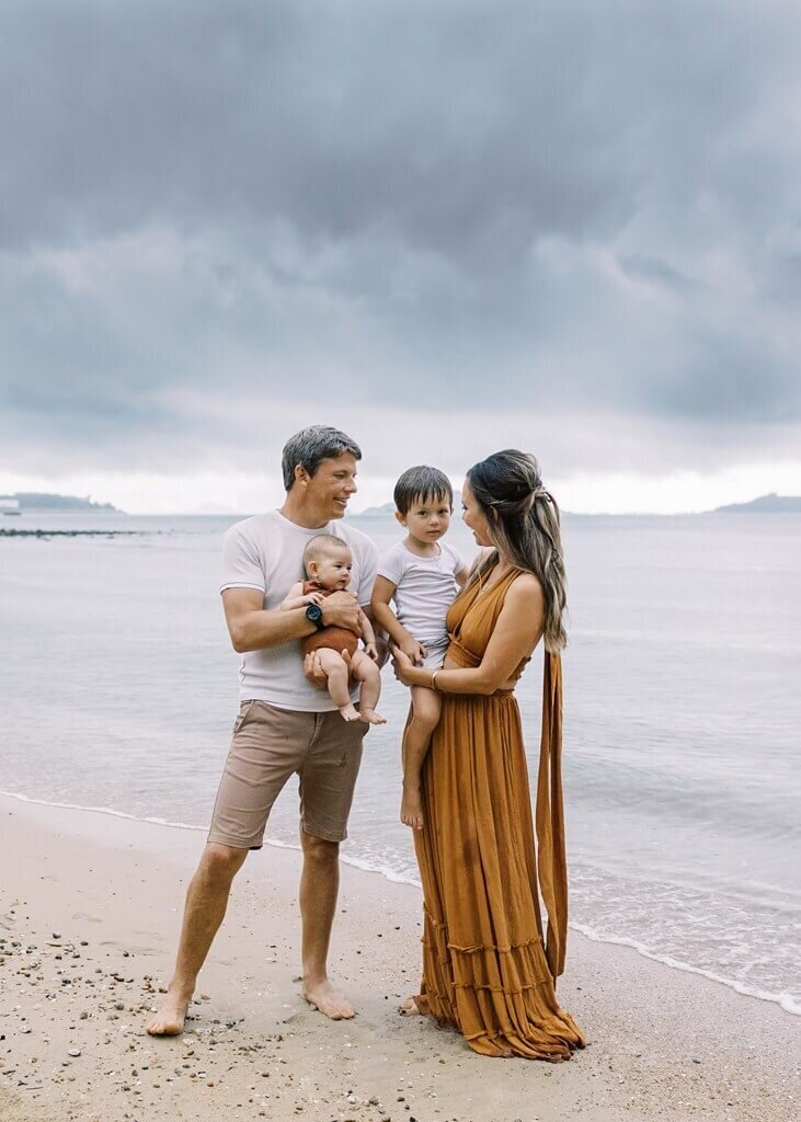 In a serene beach setting, mommy and daddy holds their children while smiling.