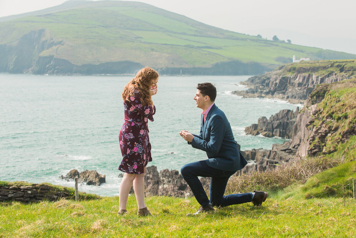 Young man going down on one knee to surprised girlfriend with red hair on a cliff overlooking the sea