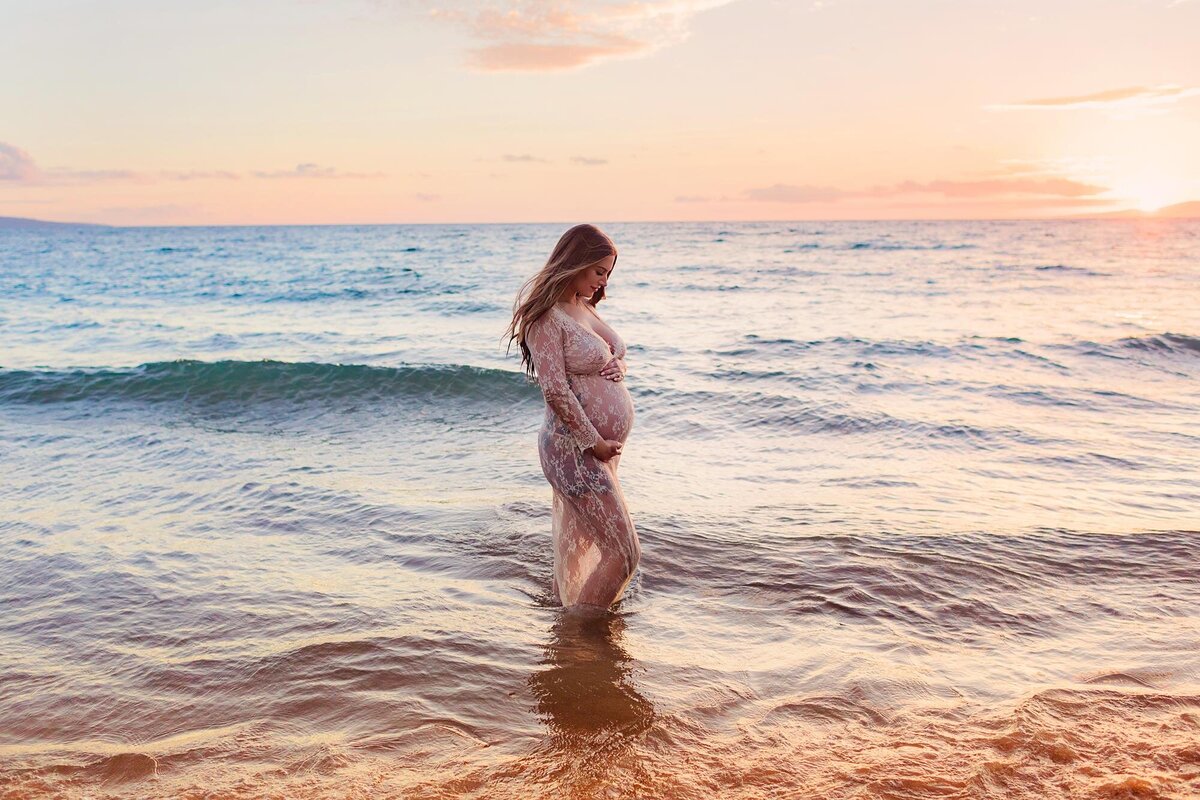 Woman in beautiful lace maternity gown holds her stomach and looks at her feet while standing in the water in Wailea