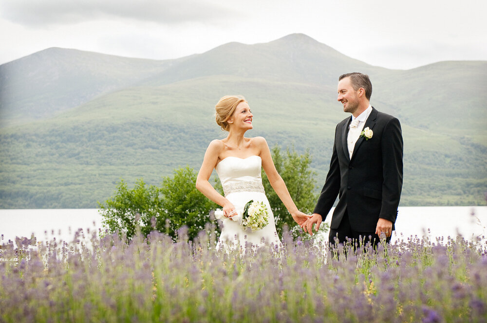red haired bride wearing an a-line dress, holding hands with her groom in a black suit and white tie surrounded by lavender flowers standing in front of a lake in Killarney