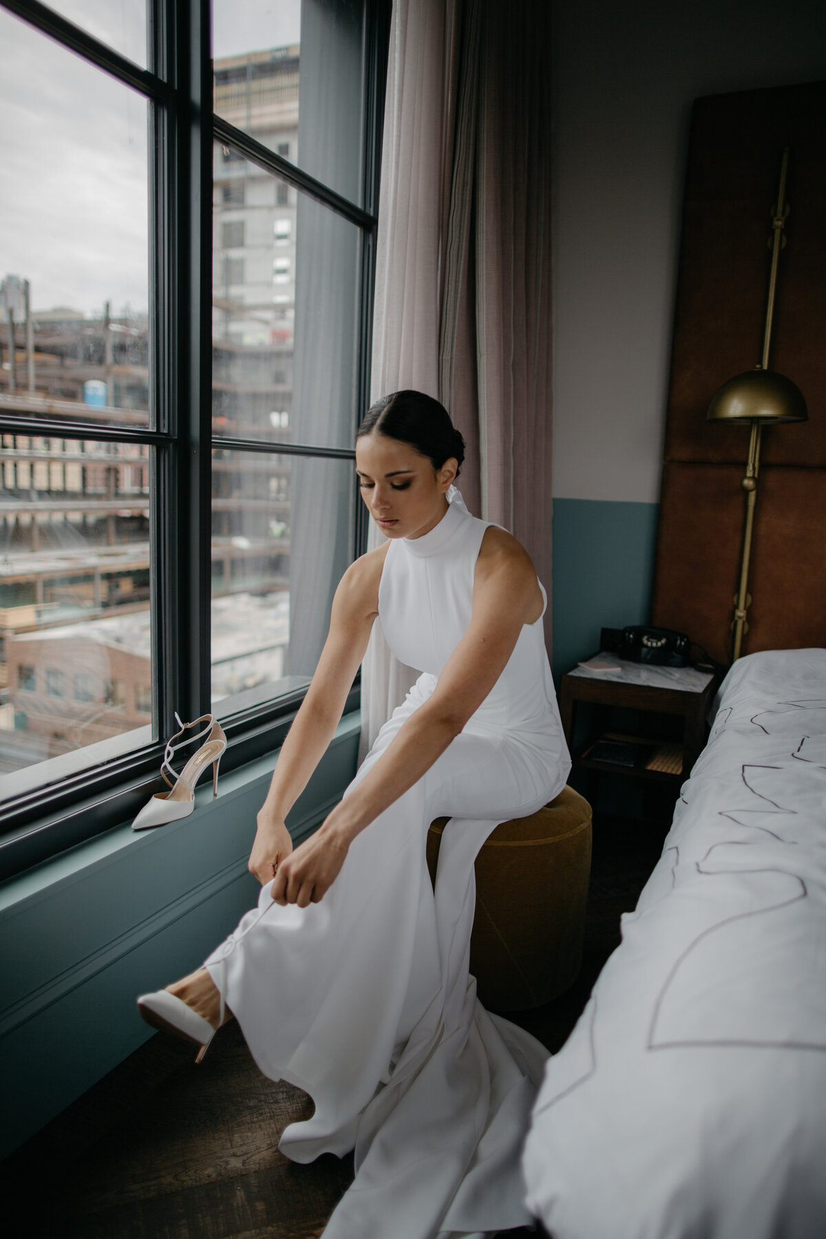 Bride sitting by window in downtown Chicago hotel room putting heels on