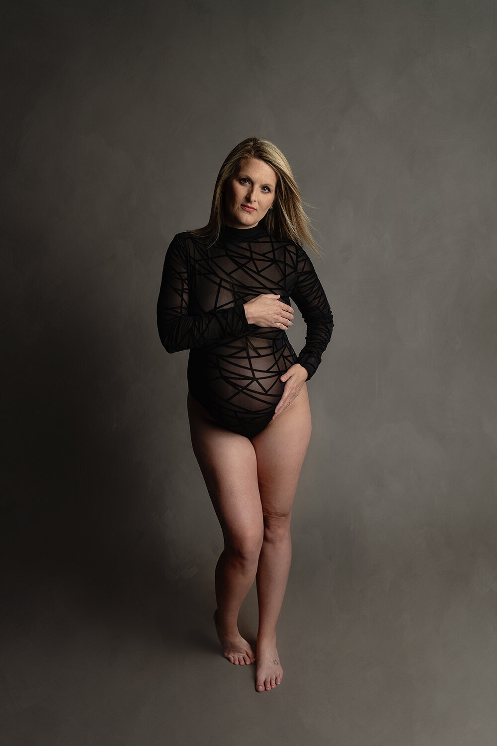 A mother to be stands in a studio with one hand on the bump in a black sheet maternity one-piece