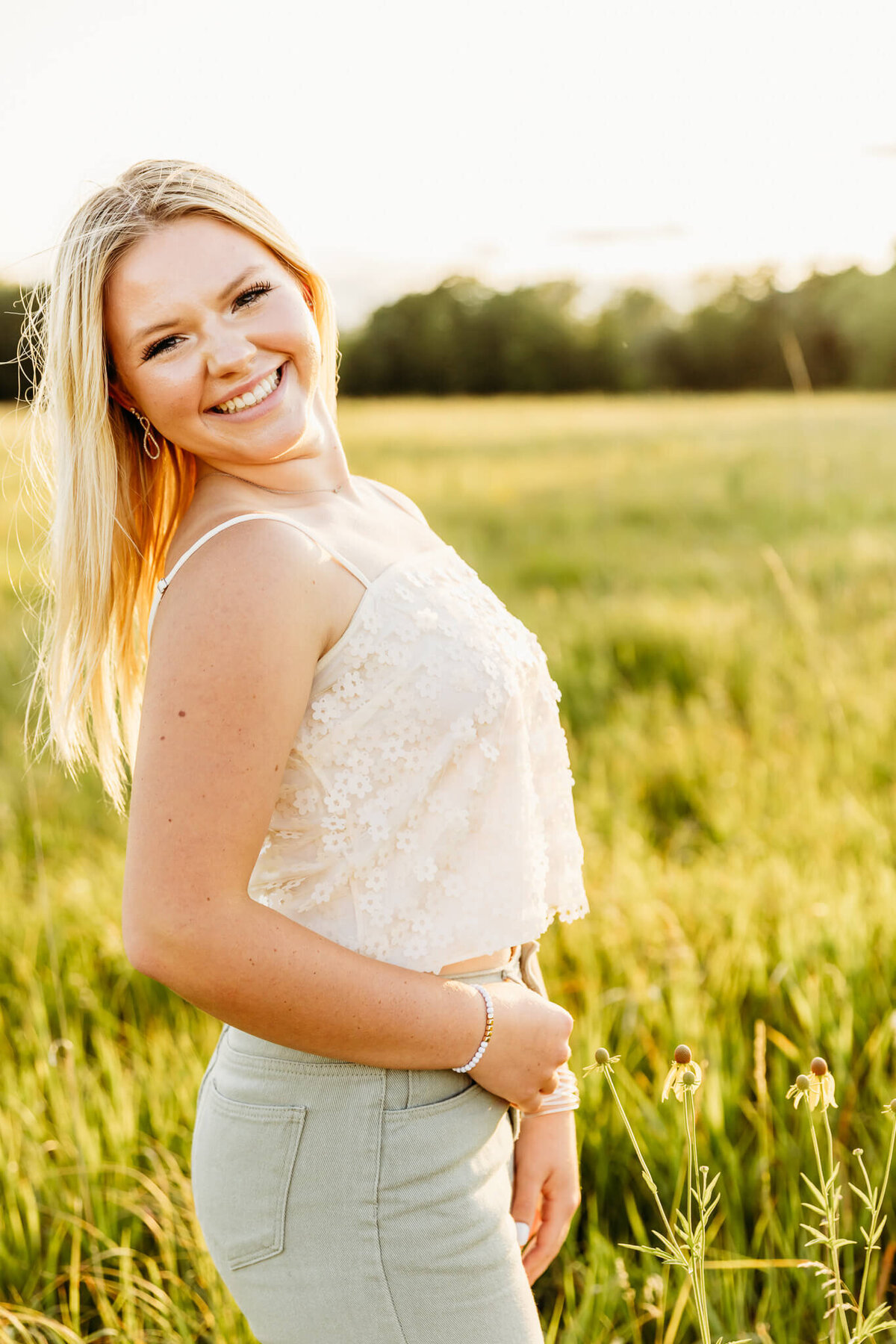 high school girl in a white flower top and green jeans looking over her shoulder in a field at sunset near Green Bay