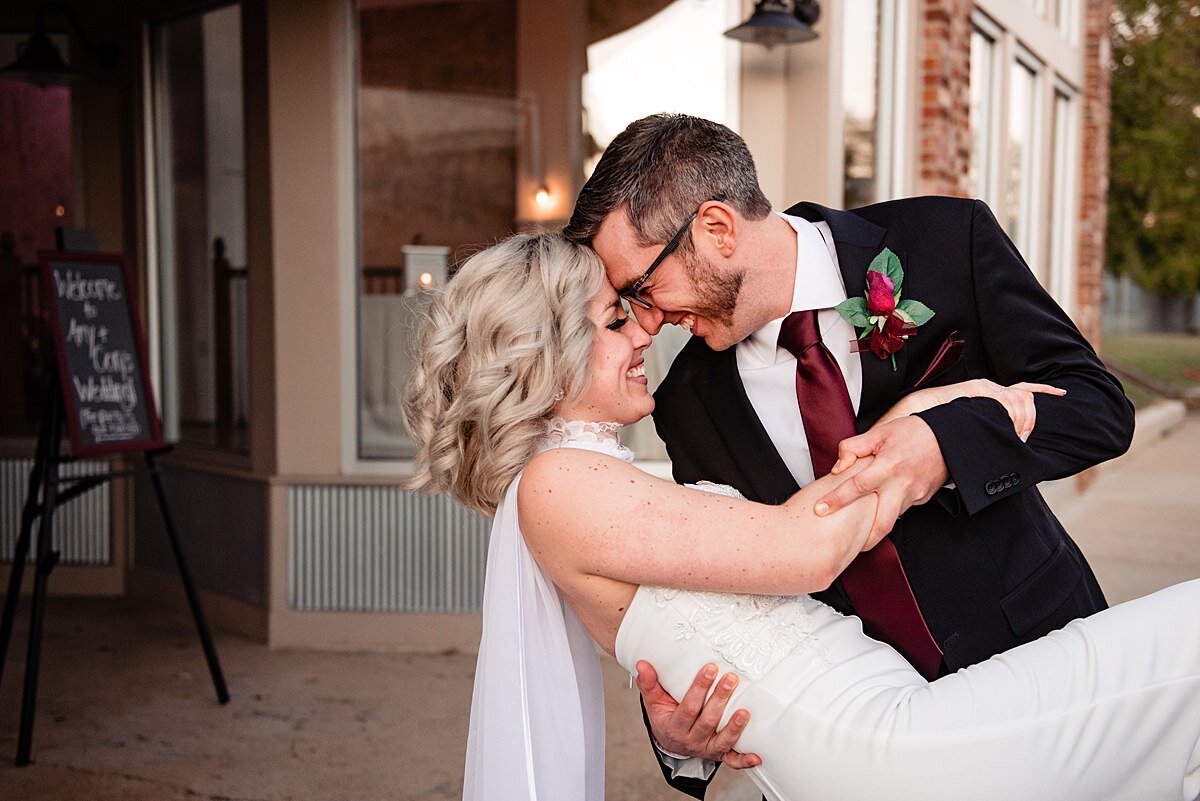 The groom dips the bride as he leans in for a kiss touching his fore head to hers. The groom is in a black suit with a deep red tie and a red rose boutonniere. The bride is wearing her hair in a low bun and her fitted white wedding dress has a long cape that comes off her shoulders.