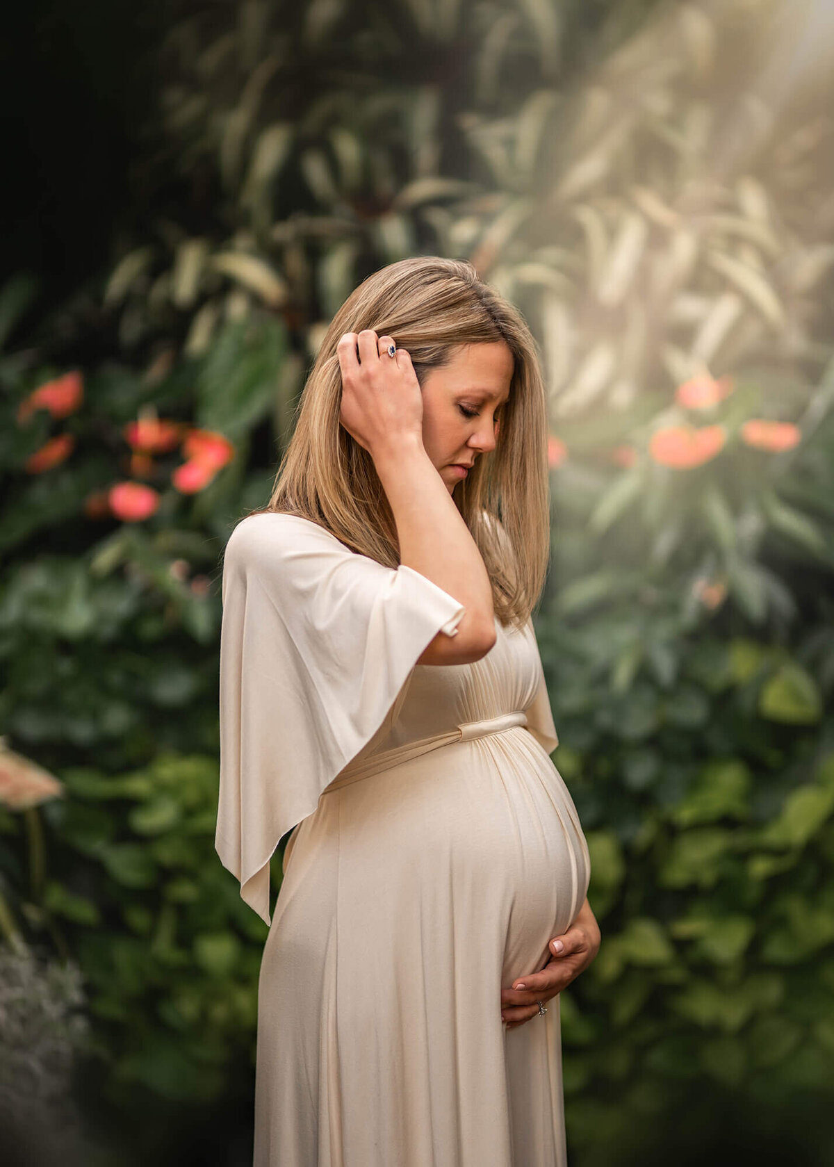 A gorgeous expecting mother in a long white dress cradles her baby bump while standing in the green house on the Biltmore Estate