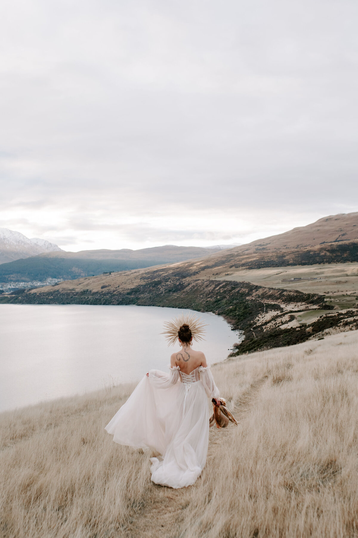 The Lovers Elopement Co - bride walks through long grass on shore of Queenstown Lake Wakatipu