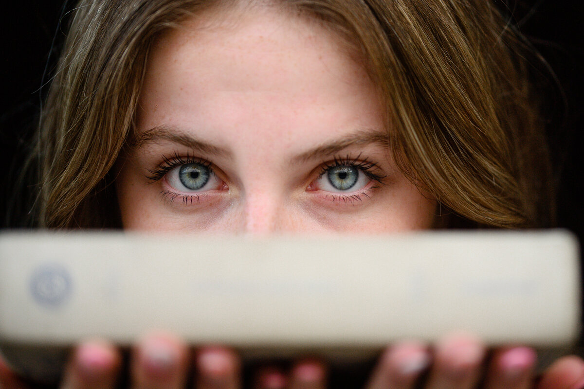 A denver senior photographer captures a closeup of a denver senior girl with bright green eyes holding a book in front of her face.