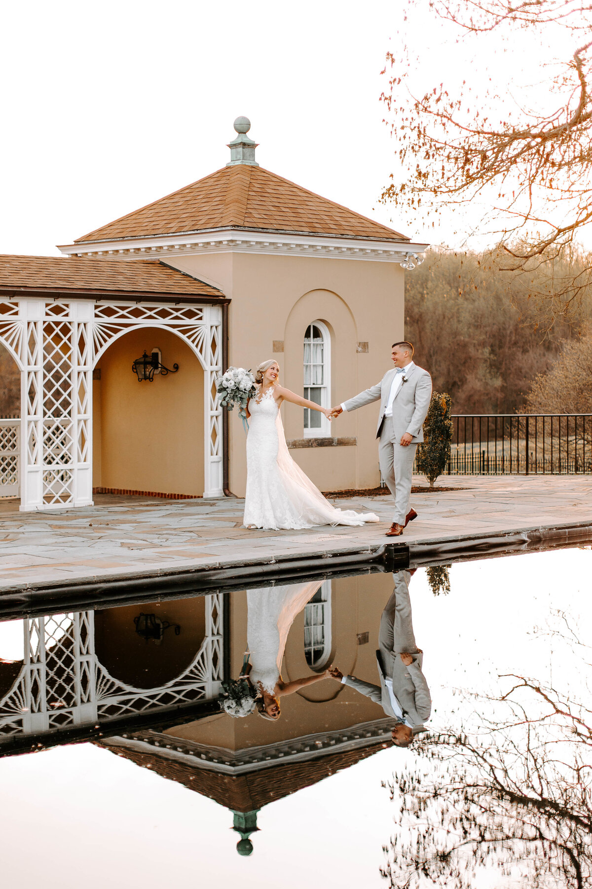 A bride and groom walk hand in hand in front of a beige building. There is a pool in front of them that perfectly reflects the image