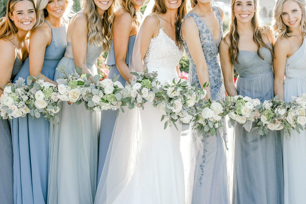 bride with bridesmaids wearing shades of light blue dresses smiling showing bouquets of white florals with greenery