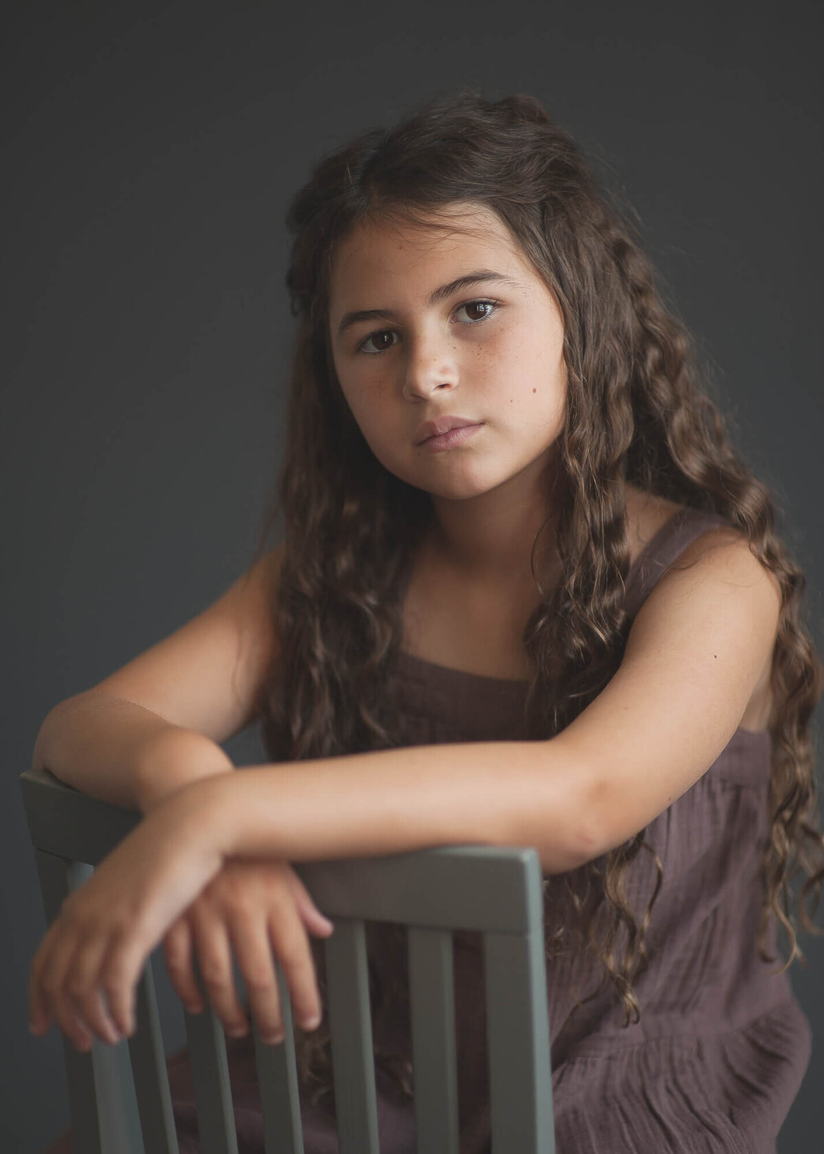 beautiful 9 year old girl with  long  curly hair studio portrait  sitting backwards on a grey chair with a dark grey background, girl is wearing eggplant color