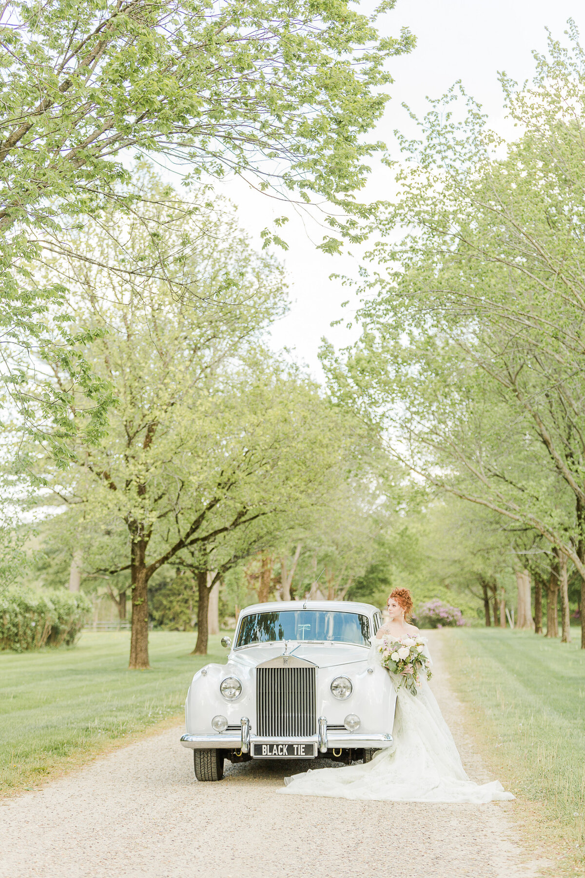 The bride leans against a white vintage Rolls Royce parked on a gravel road on an estate on Massachusetts North Shore. Captured by best Massachusetts wedding photographer Lia Rose Weddings