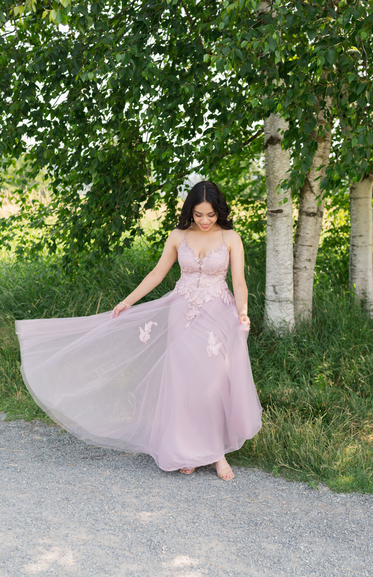 Prom day girl dressed in pink looking at her dress and swaging back and fourth, holding dress with one hand out to the side. Back round has white trees, grey trail and greenery.
