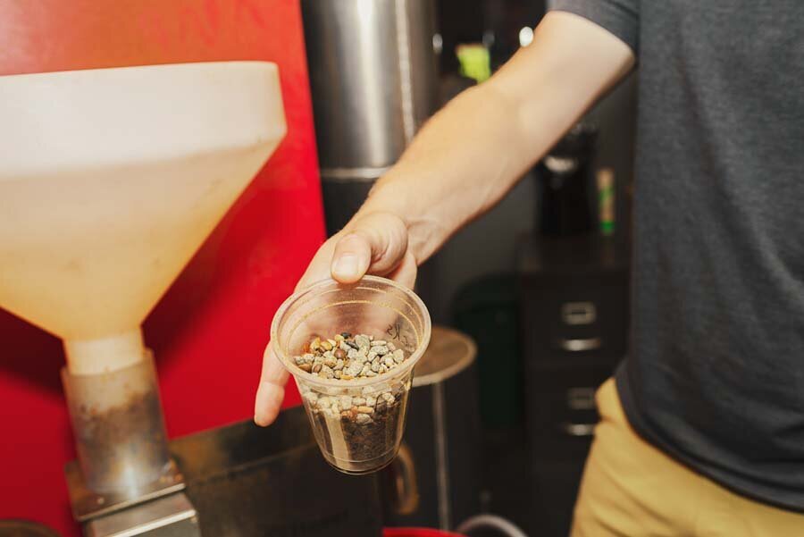 Man holding a plastic cup filled with coffee beans beside a large grinder.