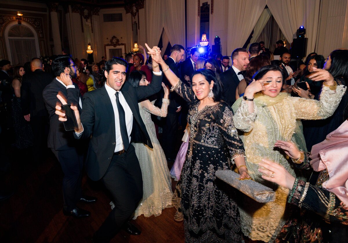 A lively crowd is dancing at an indoor event. A man in a black suit energetically moves next to women in elegant traditional attire. The room has ornate decor and soft lighting, adding to the festive atmosphere.
