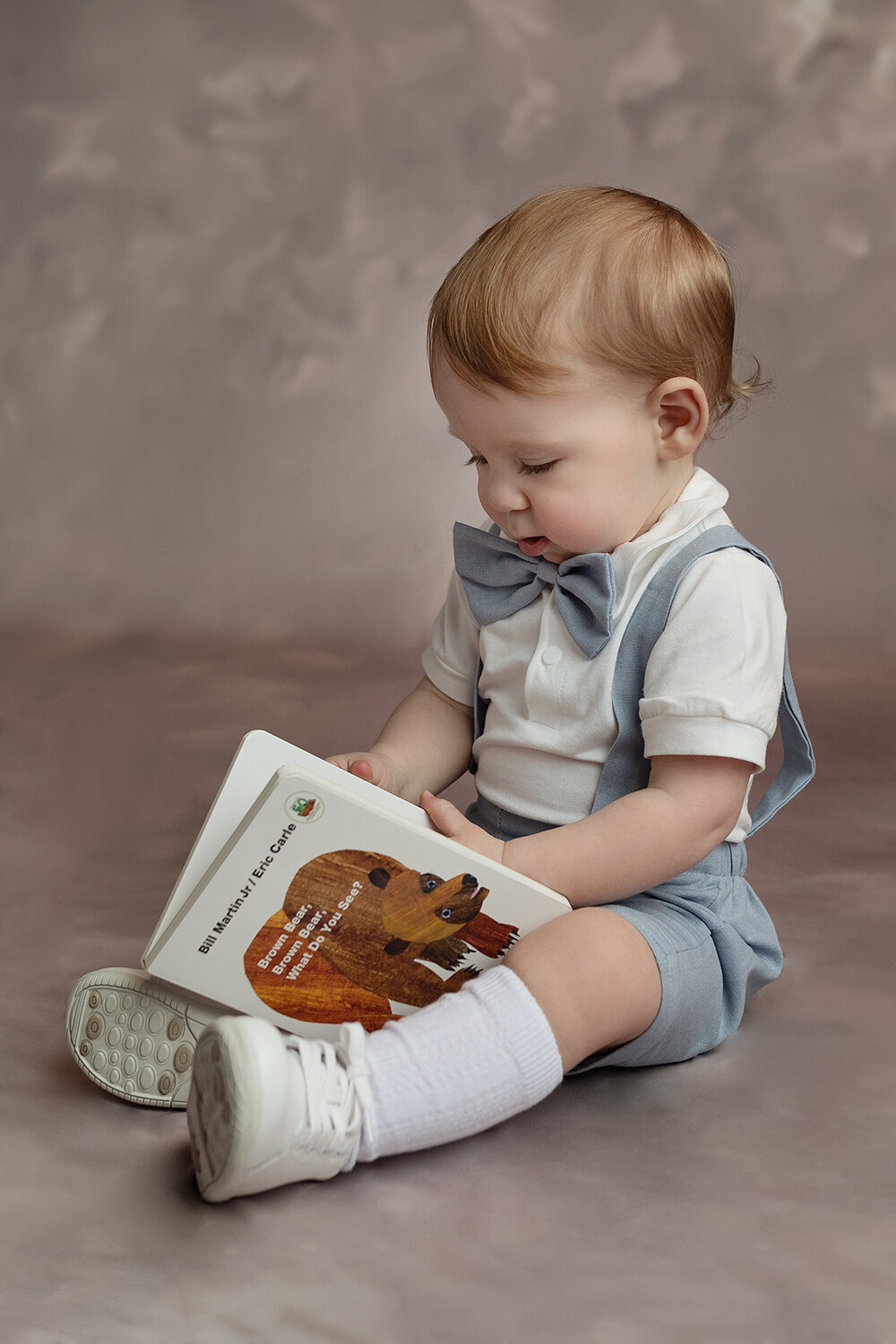 A toddler boy reads a book while sitting in a studio in blue shorts, suspenders and bowtie in a Lafayette Baby Milestone Photographer studio
