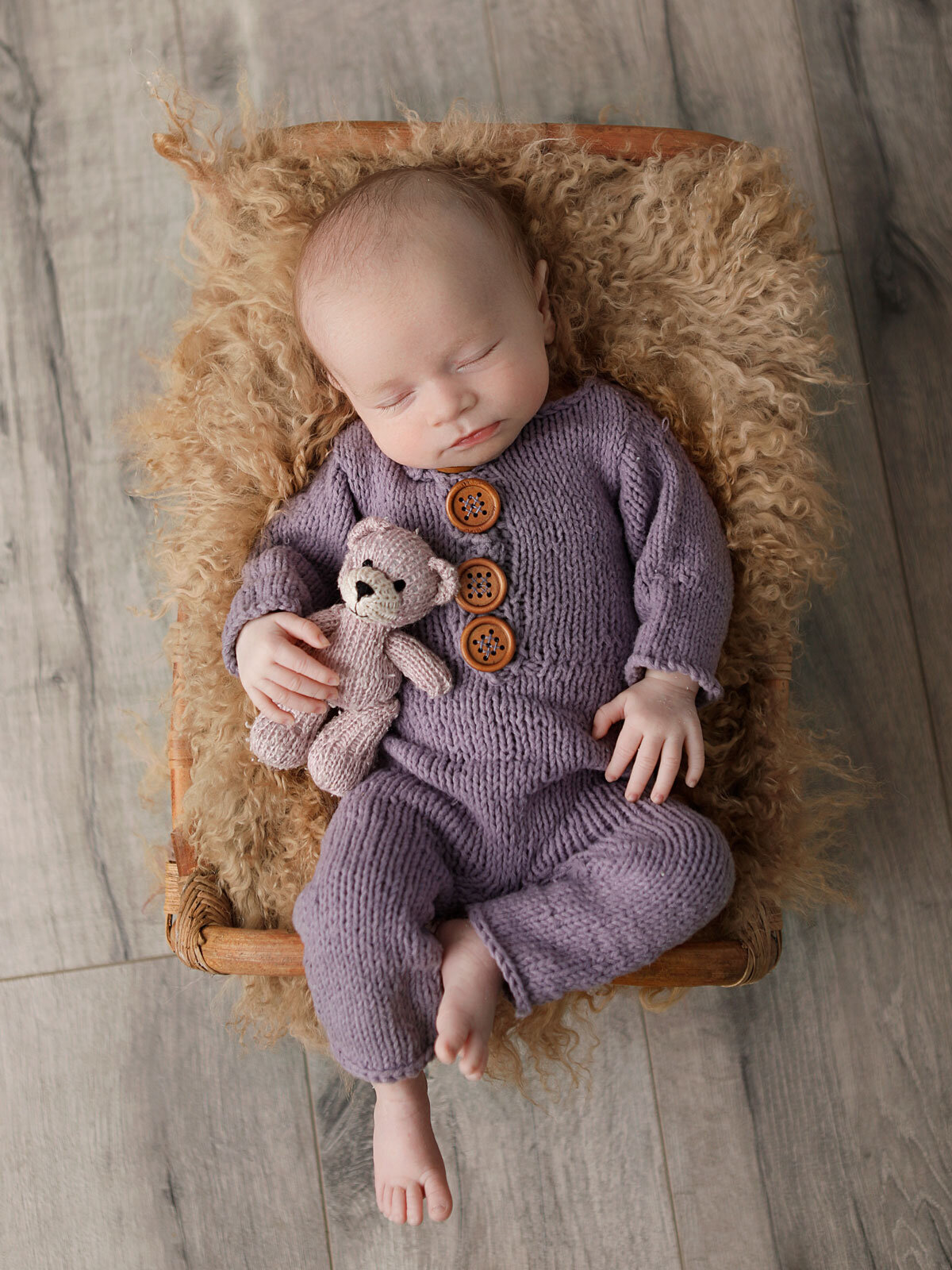 four-weeks-old-baby-girl-dressed-in-a-purple-romper-and-holding-her-teddy-bear-fast-asleep-in-her-little-bed-for-her-newborn-shoot