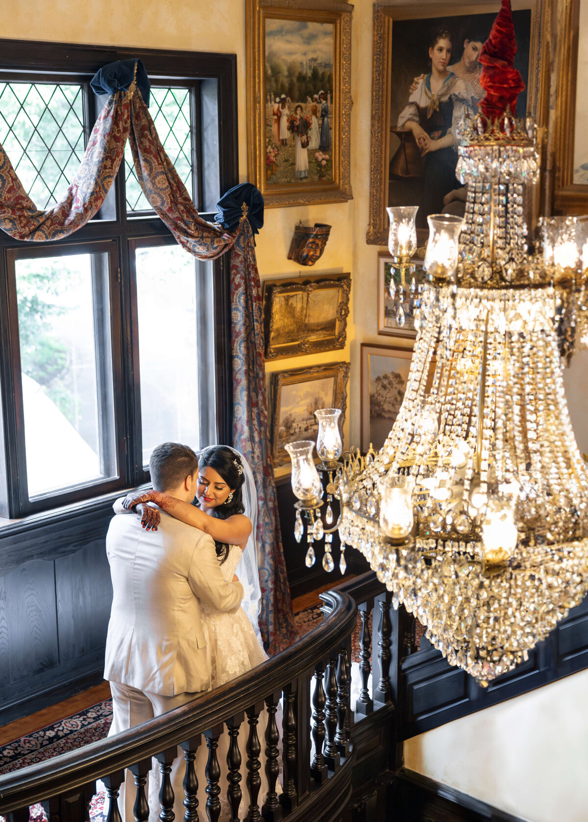 A couple embraces on a staircase in a warmly lit room adorned with paintings and a large, ornate chandelier. The room has wooden accents and intricate curtains, creating an elegant and romantic atmosphere.