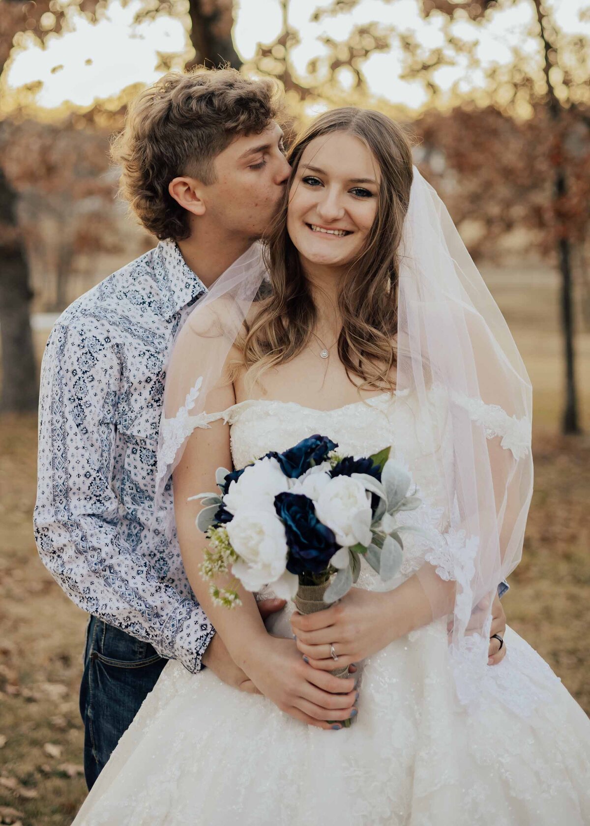 Maddie Rae Photography groom is standing behind bride with his hands on her waist. he is kissing her cheek and she is smiling at the camera holding her flowers