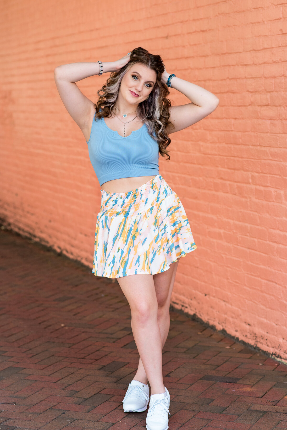 St. Catherines senior girl wearing tennis skirt and blue tank top poses with hands on head in front of coral brick wall downtown Richmond, VA.