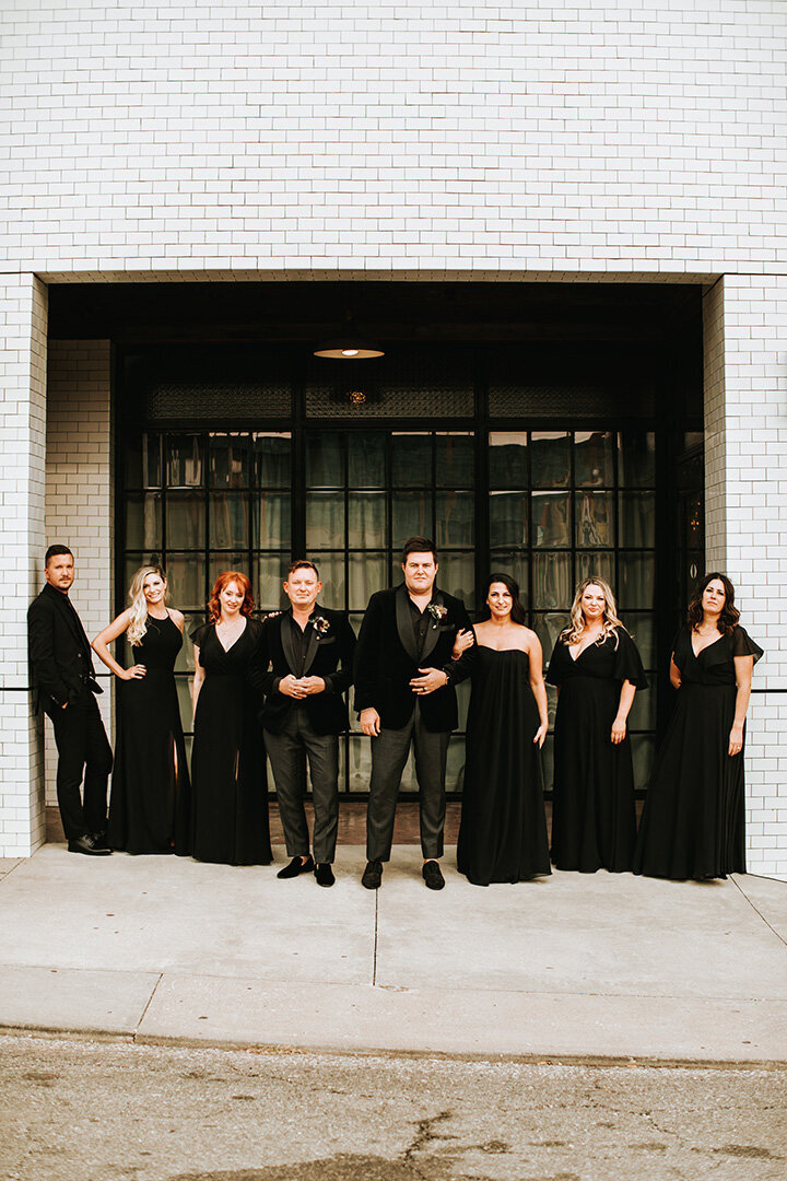 Two grooms wearing black tuxedos pose outside with their wedding party in front of a white building in the daytime.
