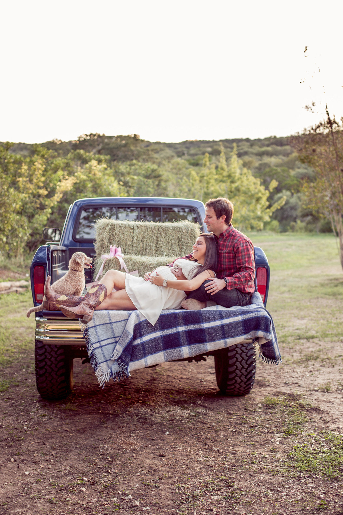 Expecting couple laying in the back of a truck with hay bales for their maternity photography session in San Antonio