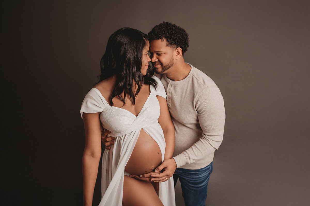 Pregnant couple standing up holding each other in maternity photo shoot wearing white sheer maternity dress with man in cream long sleeve shirt and jeans. Woman and man holding baby bump.