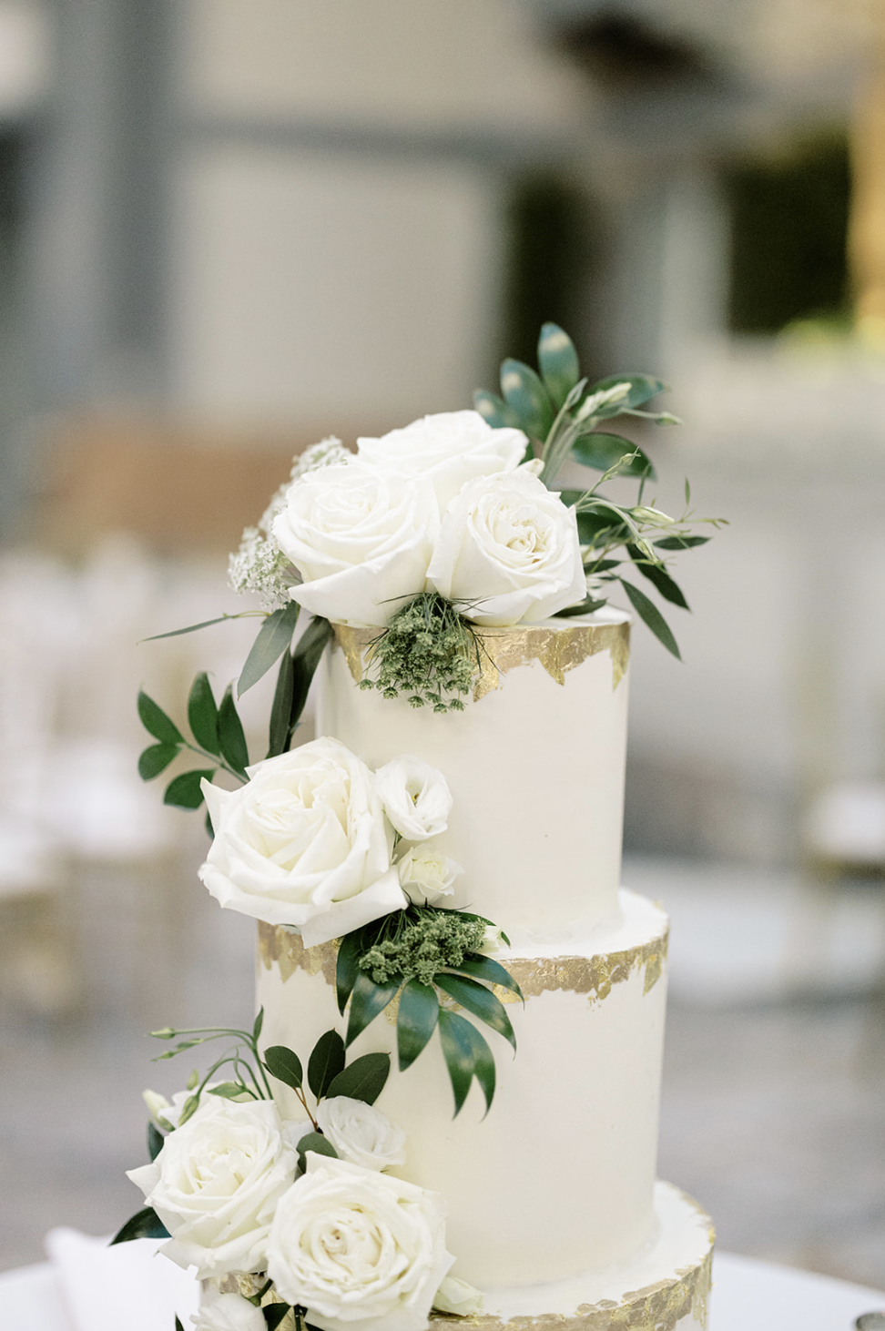 Three tier white wedding cake with gold foil outline and white roses with greenery sit on cake table at Chicago wedding reception.