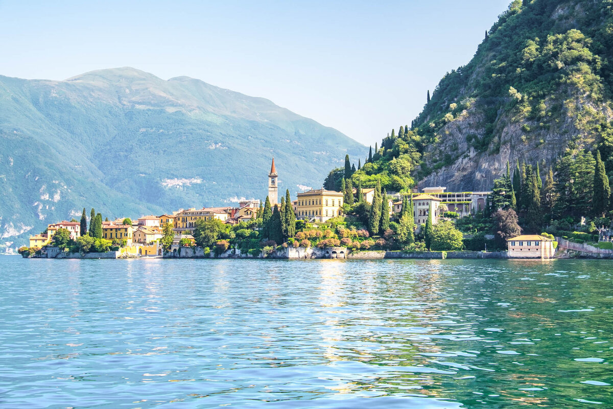 A scenic view of Lake Como with clear blue waters in the foreground and lush green hills in the background. Picturesque villas and buildings with a tall bell tower line the shoreline, surrounded by trees under a clear sky.