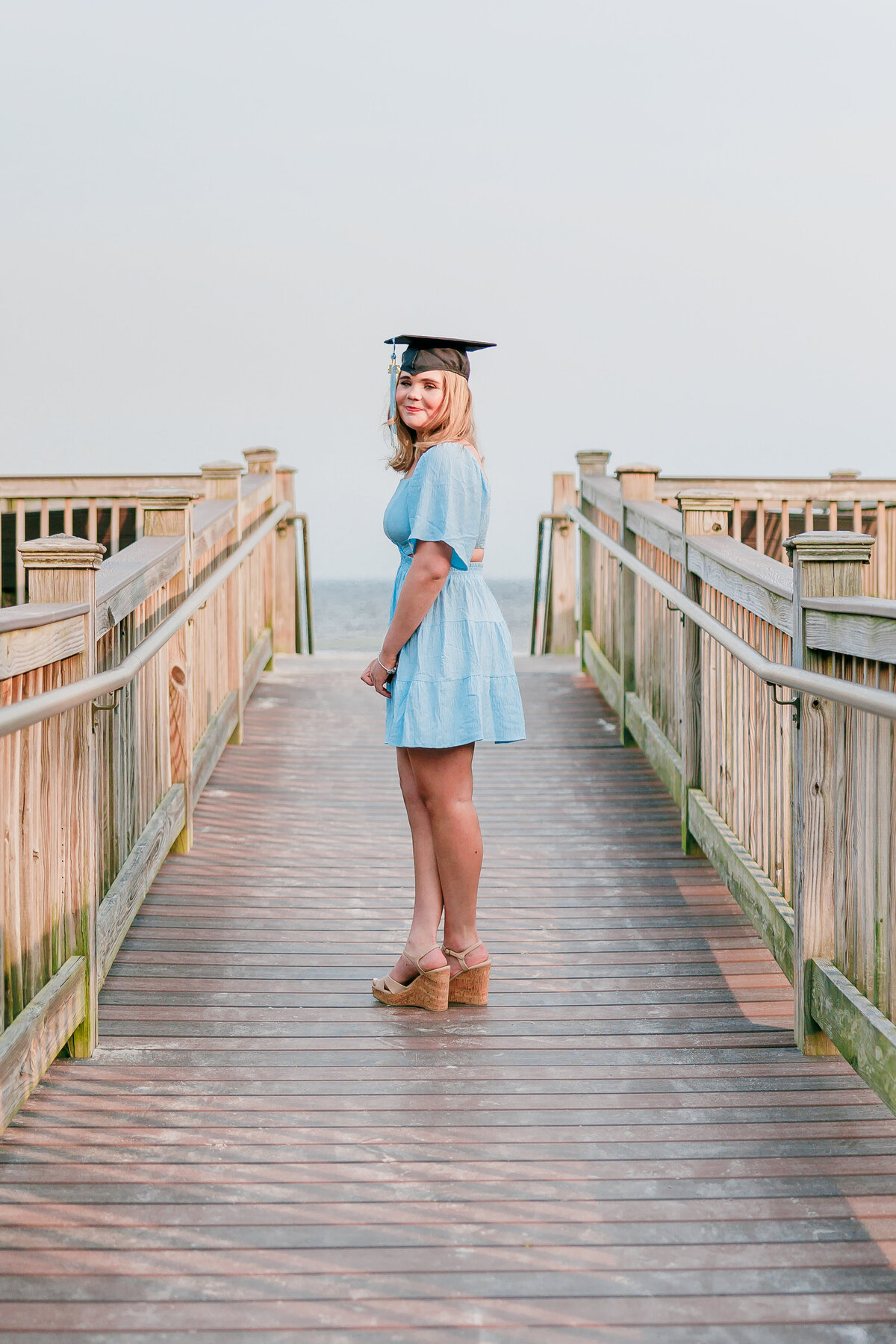 A person wearing a light blue dress and a graduation cap stands on a wooden walkway, captured perfectly by a senior photographer in Fayetteville, NC. They smile, glancing back over their shoulder, as the clear sky serves as the perfect backdrop to this memorable moment.