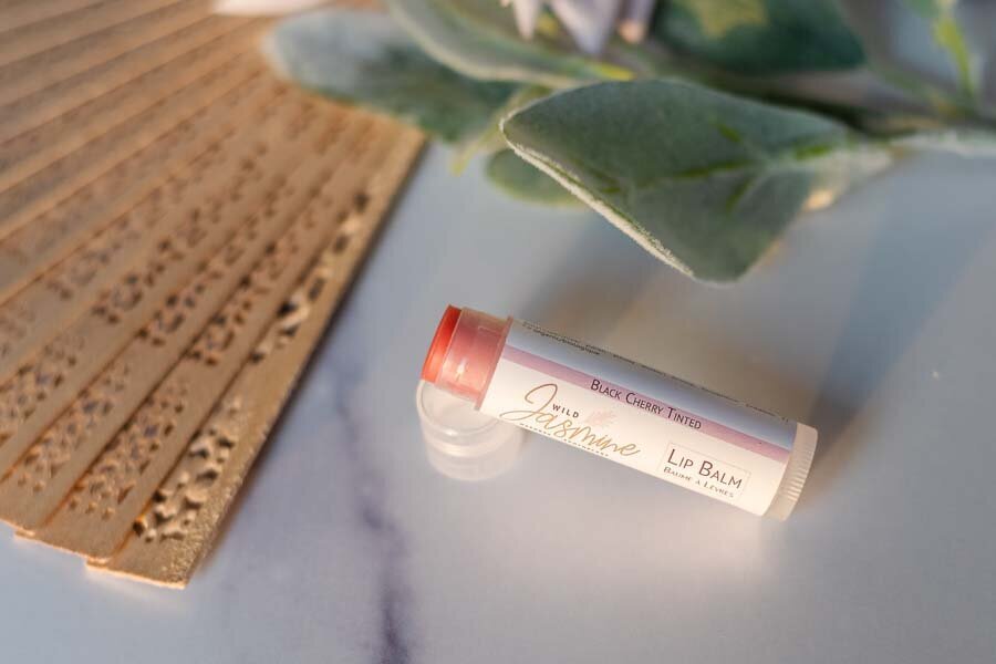 A tube of lip balm labeled "jasmine" beside green leaves on a marble surface, with a bamboo mat partially visible in the background.