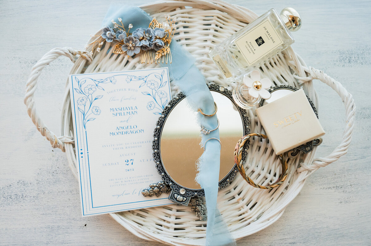 A white wicker tray with various wedding details like a small mirror, invitation, perfume bottle, jewelry and blue ribbon.