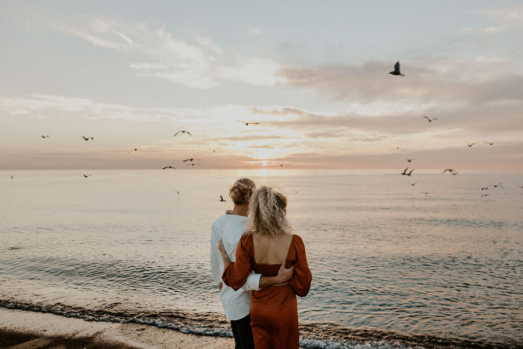 Man with a manbun wearing a white shirt hugging woman in red dress while they look out over the ocean