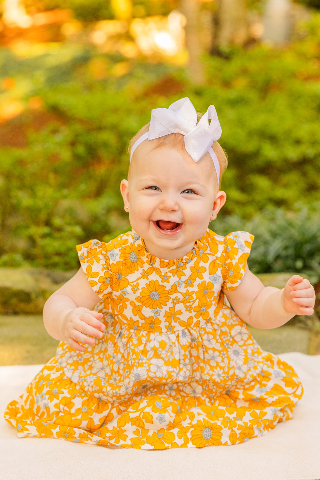 Baby one year girl wearing a yellow dress with flowers sitting on a white blanket and smiling at the camera on a Atlanta park path by Laure Photography