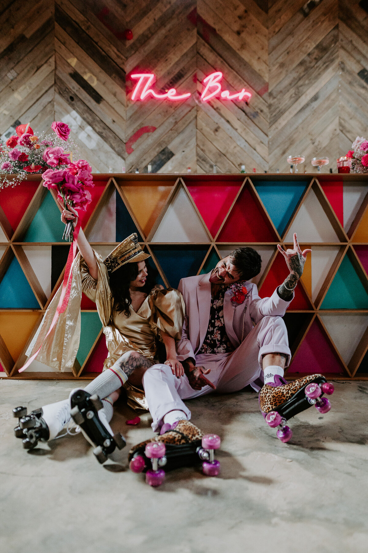 A couple wearing roller skates at their wedding at The Canary Shed. The groom is wearing a lilac suit and the bride is wearing a golden dress, they are both heavily tattooed and alternative. The bride is holding up a pink wedding bouquet which matches the colour of their wedding.