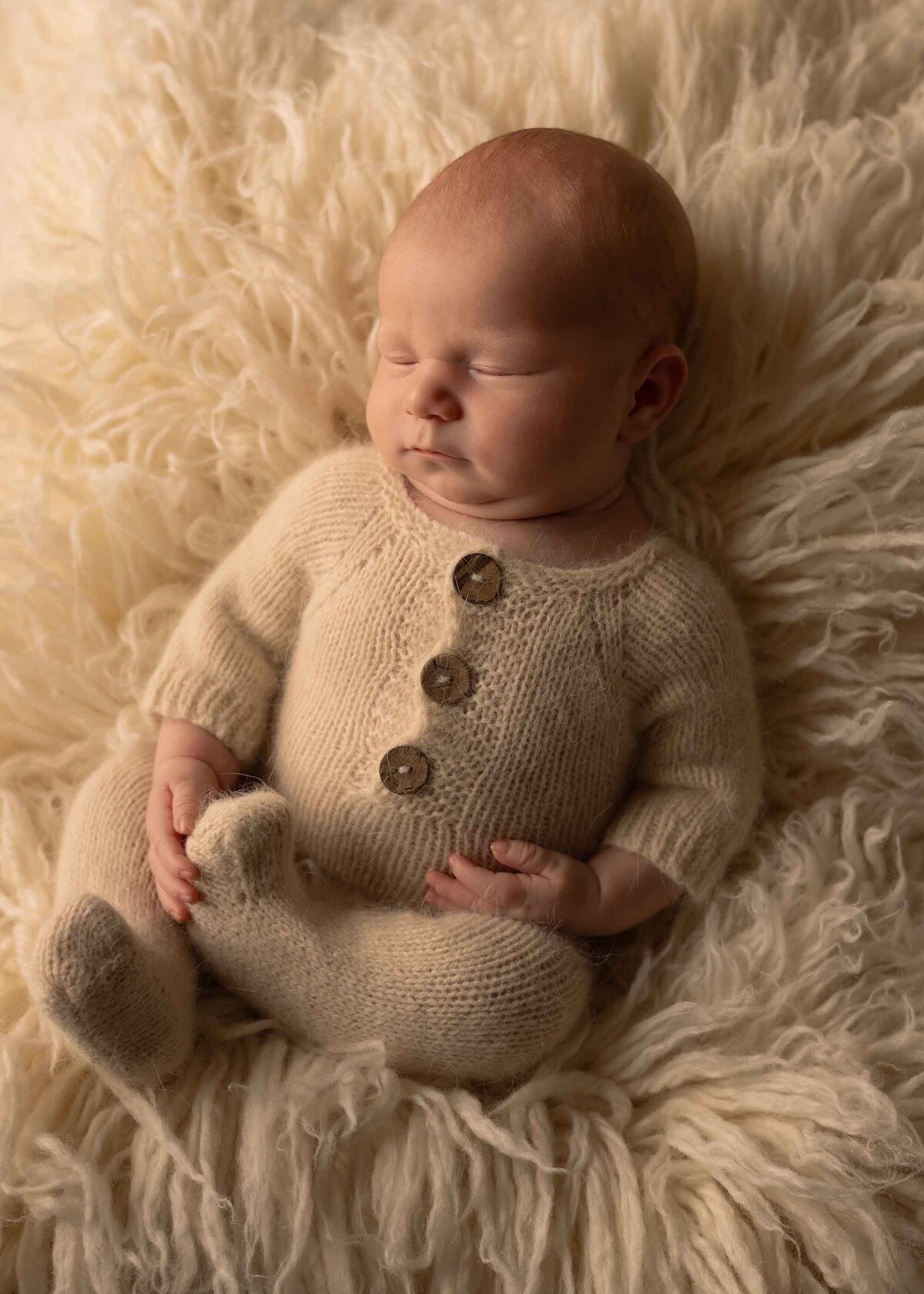 Maddie Rae Photography off to the side angle of baby sleeping with his legs crossed while laying on a fluffy rug. he is wearing a neutral colored outfit