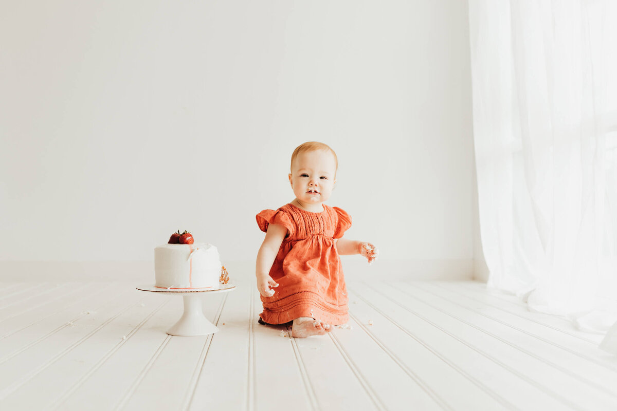 Little girl kneeling on a white wood floor with her cake on an all white backdrop by a window.