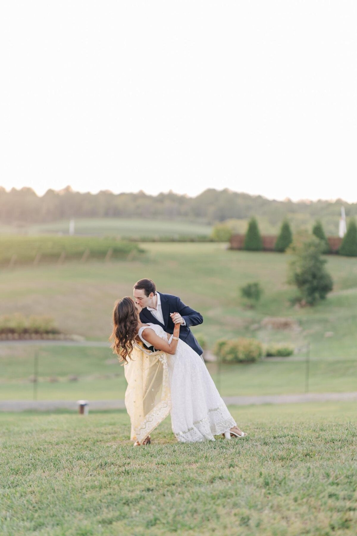 A couple in wedding attire kissing, with the groom dipping the bride in a grassy field, gentle hills in the background.