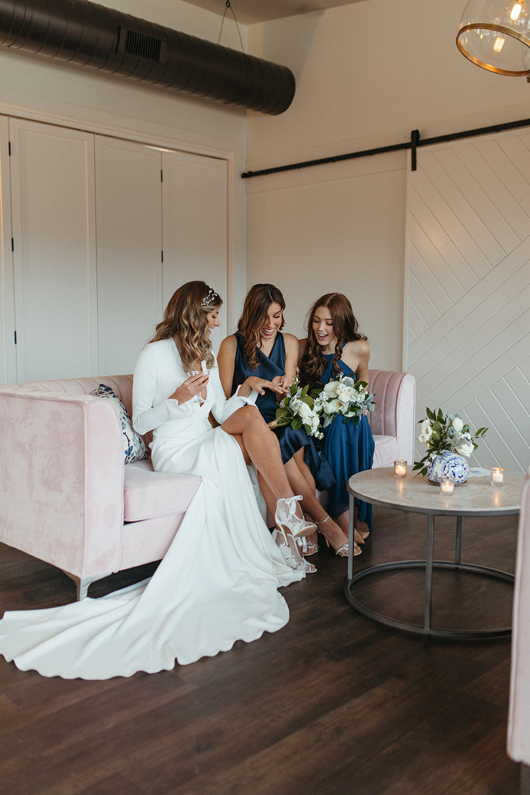 Bride in a white wedding gown sits with two bridesmaids in blue on a light pink couch.