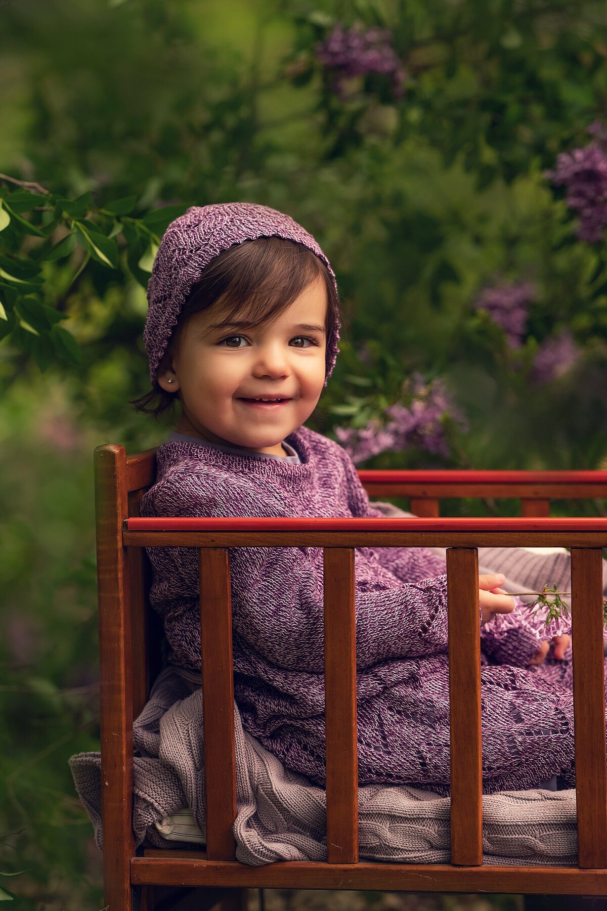 A young child sits outdoors in vintage crib surrounded by purple flowers for a whimsical, stylized portrait.