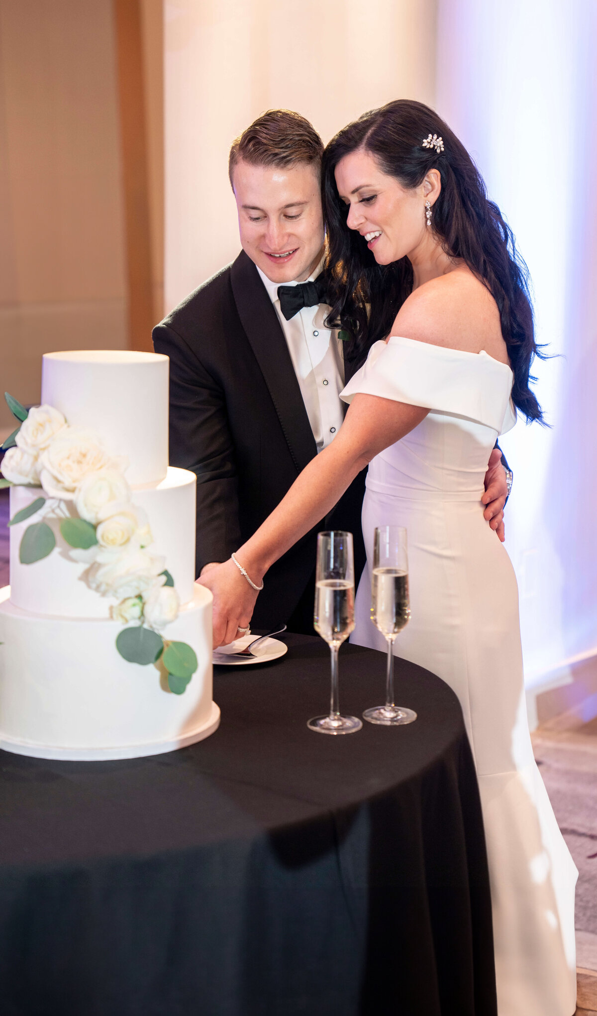 A couple in formal attire is cutting a three-tiered white wedding cake adorned with white roses. Two champagne glasses are on the table. The bride wears a white off-the-shoulder dress, and the groom is in a black tuxedo.
