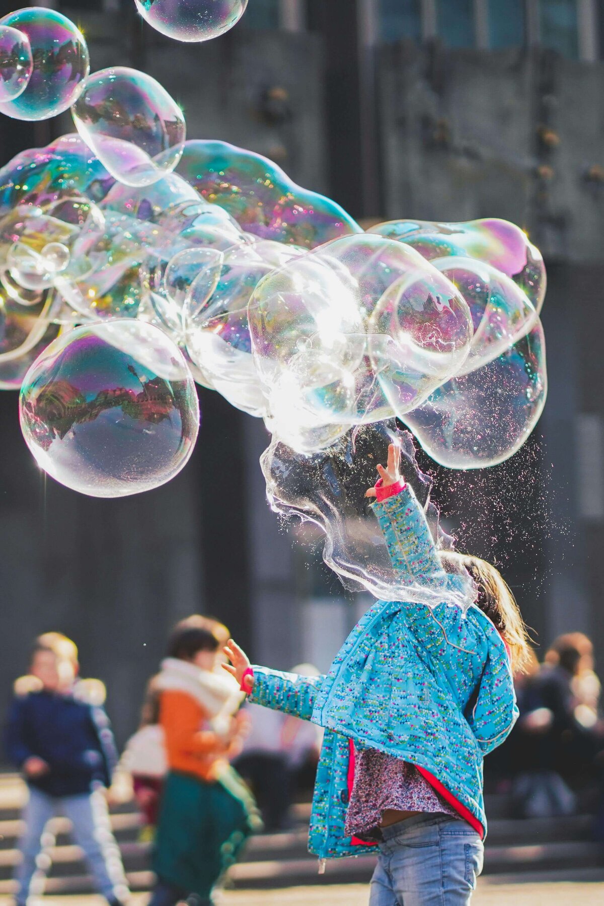 A child in a bright blue jacket reaching up to pop large, iridescent bubbles in an outdoor setting, surrounded by other children playing in the background.