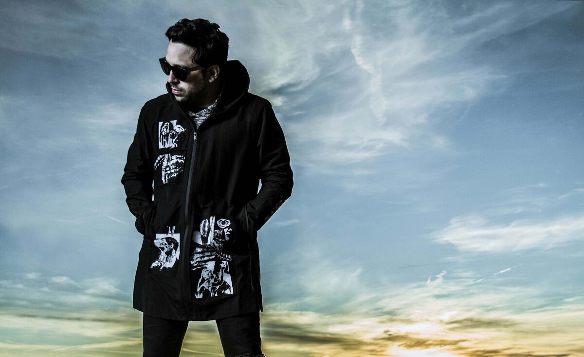 Male musician portrait Dagg3rs wearing black coat with white writing standing against desert sunset background