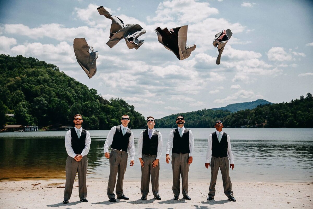 Groomsmen on a lakeside beach tossing their jackets into the air, with a scenic mountain and lake in the background.