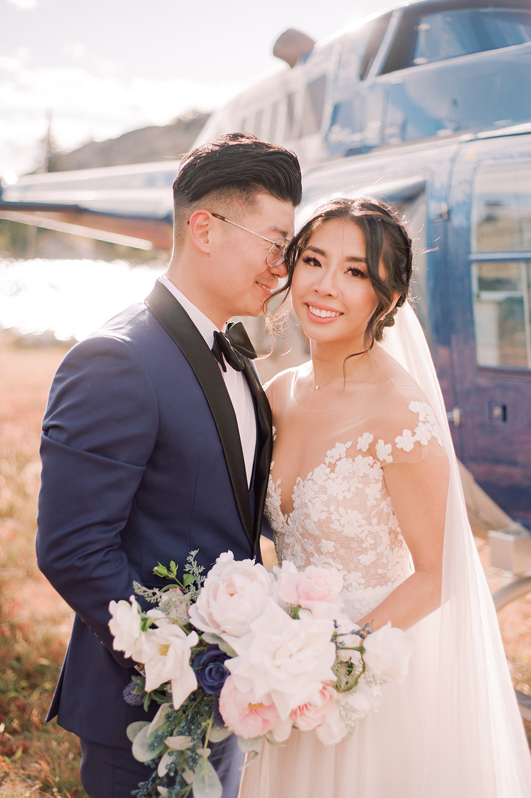 Newlyweds  stand in front of a blue helicopter on the top of a mountain and smile.