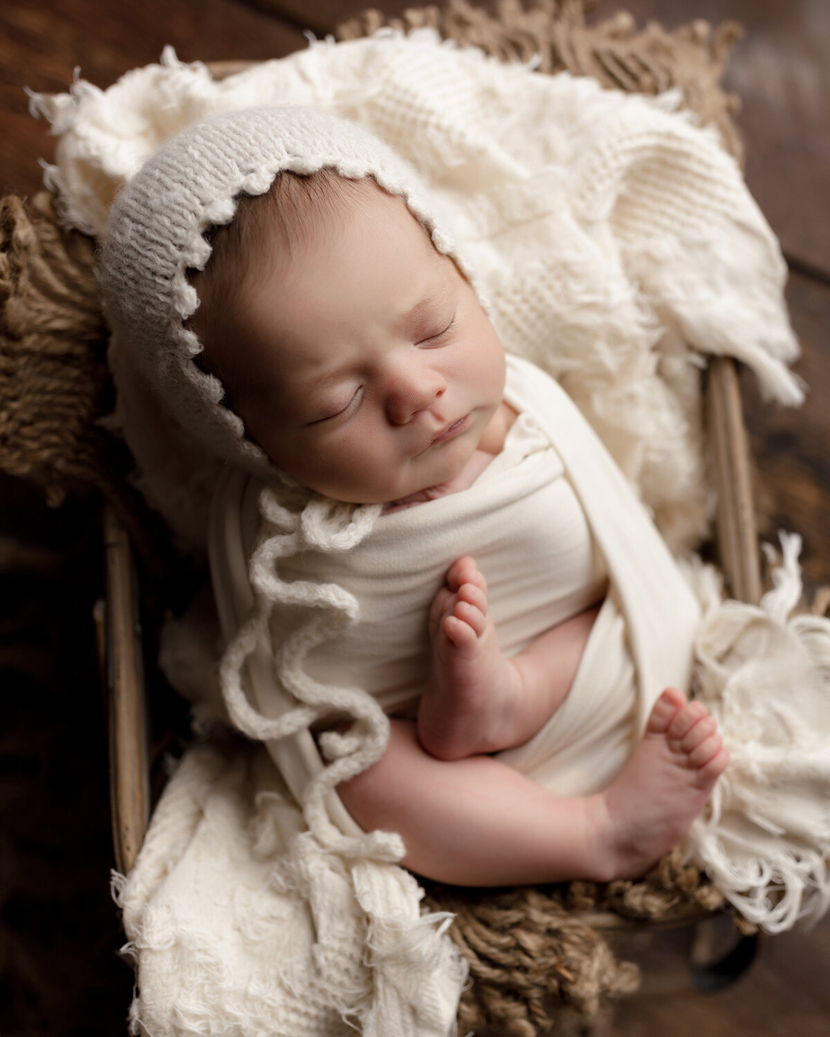 Newborn Photos in top West Palm Beach Photography Studio - Baby girl is wrapped in a cream coloured stretchy swaddle with her legs exposed and posed. She is wearing a cream coloured knit bonnet and sleeping atop layer of plush cream and tan blankets in a crate. Aerial image.