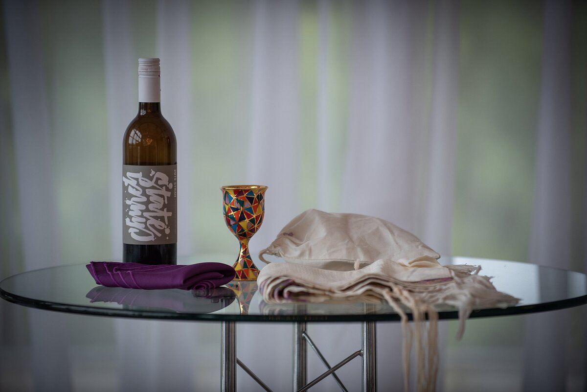 On a tall glass table sits a bottle of wine with a purple napkin and a gold enameled  kiddush cup next to the Jewish groom's tallit.