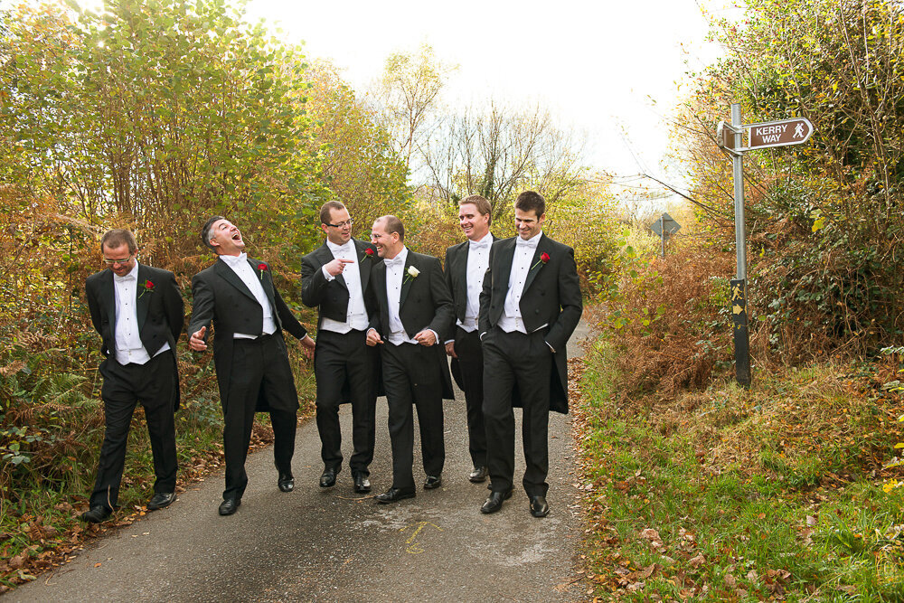 Groom and groomsmen in white tie and black tailcoats walking together on country road laughing